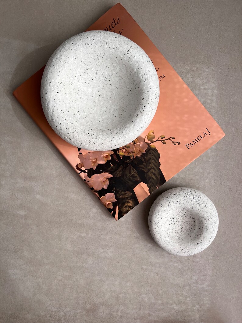 May include: Two white speckled ceramic bowls, one larger than the other, on a pink and white book with the text "Bouquets" and "Pamela J".