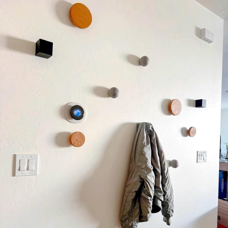 May include: A white wall with a variety of wall hooks, including round wooden hooks, round grey hooks, and a square black hook. A grey jacket is hanging on one of the hooks. There is a light switch on the wall.