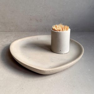 May include: A white concrete tray with a rounded edge and a white concrete toothpick holder with wooden toothpicks.