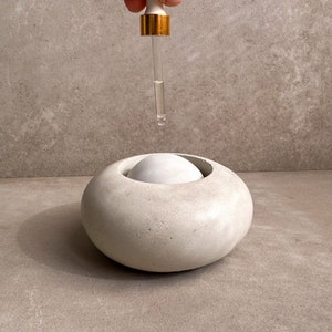 Może przedstawiać: Biały betonowy dyfuzor olejków eterycznych o okrągłym kształcie z białym kamieniem w środku. Pipeta ze złotą końcówką dozuje płyn do dyfuzora. Tło jest neutralnie szare.