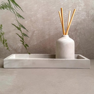 May include: A gray concrete tray with a white ceramic diffuser bottle and natural reed sticks. The diffuser bottle is sitting on the tray. The tray is on a gray surface.