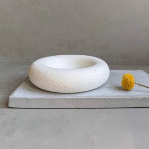 May include: White speckled concrete ring dish with a small yellow flower on a grey concrete surface.