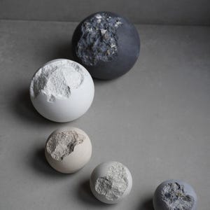 Puede incluir: Colecci&oacute;n de objetos decorativos esf&eacute;ricos de varios tama&ntilde;os y colores. Cada esfera tiene una secci&oacute;n de su superficie eliminada, revelando un interior texturizado y contrastante. Los colores incluyen blanco, gris y un beige apagado.