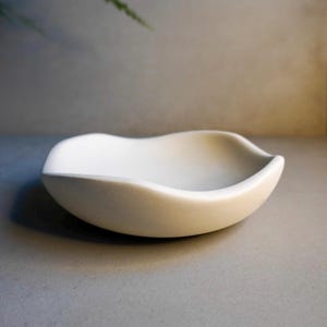 Puede incluir: Un pequeño cuenco de cerámica blanquecina con un diseño suave y curvado. El cuenco tiene poca profundidad y un borde ligeramente irregular, lo que le da una forma orgánica única. El color neutro y el estilo minimalista lo convierten en una pieza versátil.