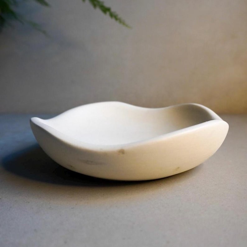 May include: A small, cream-coloured, shallow bowl with a distinctive, curved form. The bowl has a smooth surface, possibly ceramic or stone. The design is simple and elegant, suitable for serving small portions or as a decorative piece.