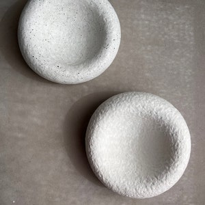 May include: Two white ceramic bowls with a textured surface. The bowls have a rounded shape and a slight indentation in the center.