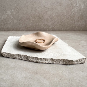 May include: A light brown, wavy ceramic dish with a gold ring inside, sitting on a light gray, textured stone surface.