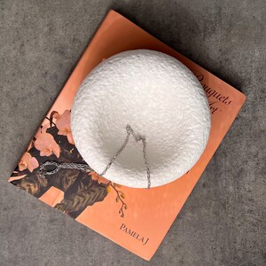 May include: White round hanging planter with a silver chain. The planter is sitting on a book with the text "Bouquets of Art" and "Pamela J" on the cover.