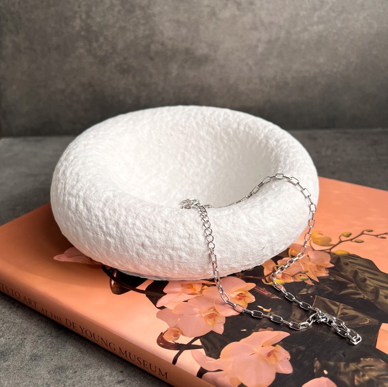 May include: A white ceramic bowl with a textured surface, designed to hold jewelry. A silver chain necklace is resting inside the bowl.
