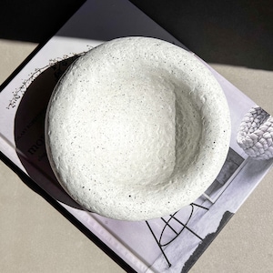 May include: A white speckled ceramic bowl with a rounded rim. The bowl is sitting on a book with a black and white image of a chair.