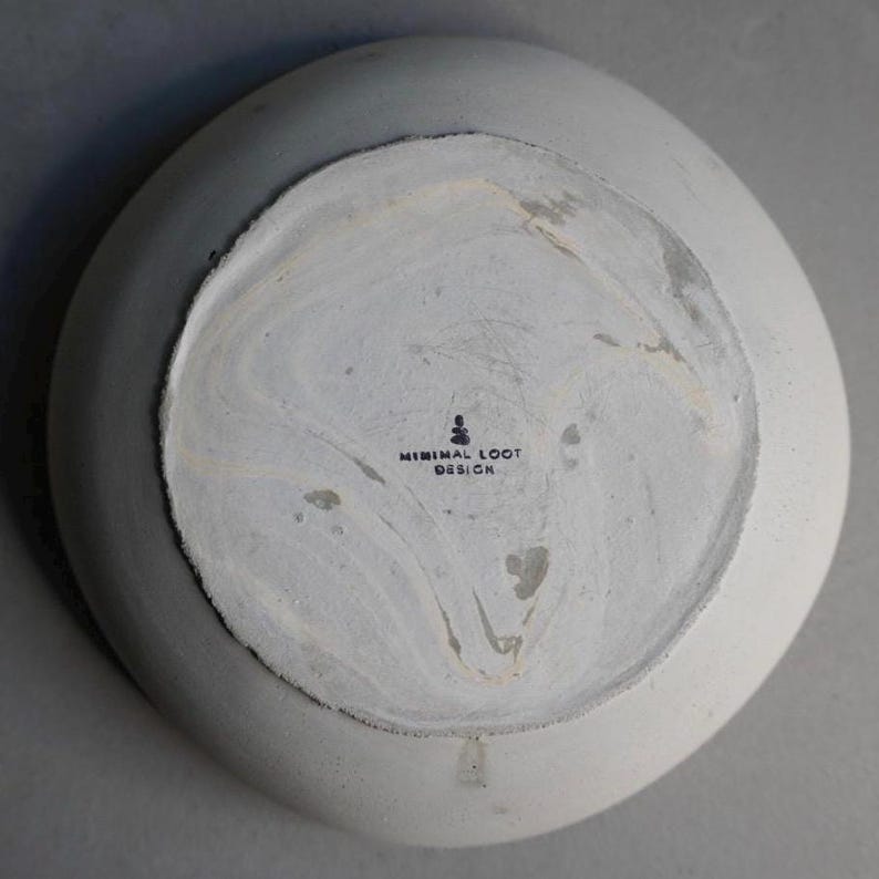 May include: A light grey, round ceramic bowl with an irregular shape. The base of the bowl has a marbled white and grey pattern and the text "MINIMAL LOOT DESIGN".