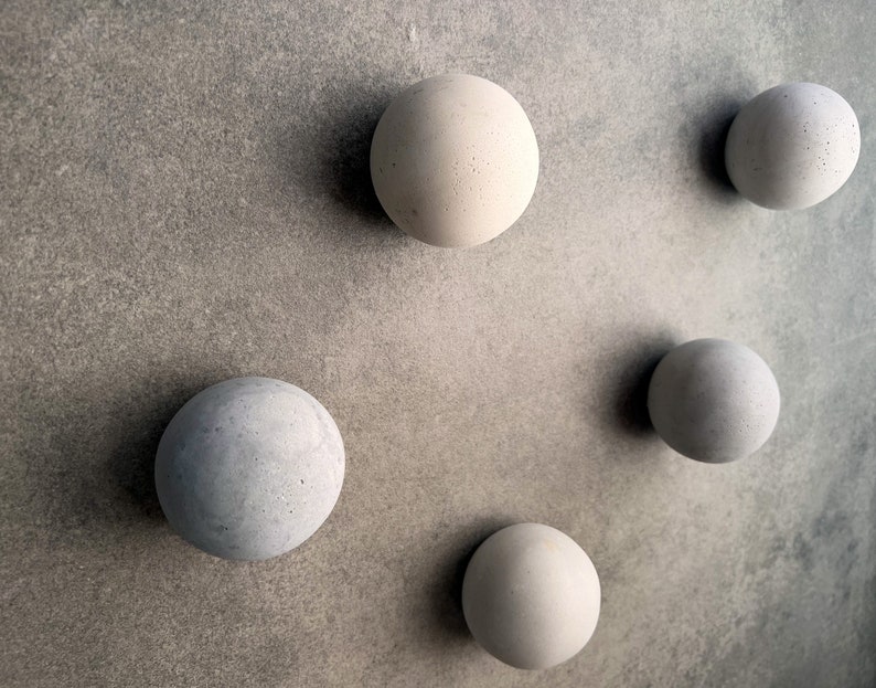May include: Five round, gray concrete spheres of varying sizes are arranged on a gray textured surface.