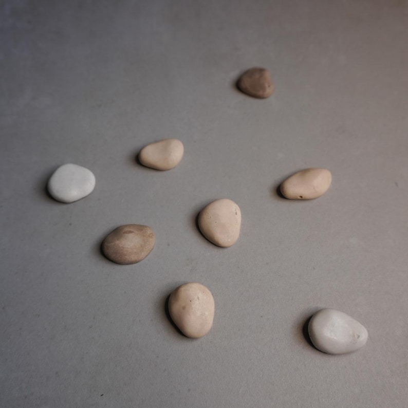 K&ouml;nnte beinhalten: Eine Auswahl an glatten, abgerundeten Steinen in verschiedenen Beige- und Braunt&ouml;nen, mit einem wei&szlig;en Stein. Die Steine sind auf einer grauen Oberfl&auml;che angeordnet und erzeugen eine nat&uuml;rliche, minimalistische &Auml;sthetik. Ideal f&uuml;r Heimdekoration oder Bastelprojekte.