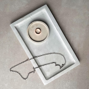 May include: A gray concrete tray with a small round indentation and a silver chain necklace. The indentation has a gold ring inside.