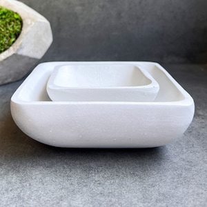 May include: Two white square ceramic bowls, one nested inside the other. The bowls have a smooth, slightly textured surface.