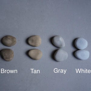 K&ouml;nnte beinhalten: Vier Paare glatter, abgerundeter Steine in Braun, Beige, Grau und Wei&szlig;, angeordnet auf einer grauen Oberfl&auml;che. Die Farben sind unter jedem Paar beschriftet. Die Steine haben ein nat&uuml;rliches, organisches Aussehen.