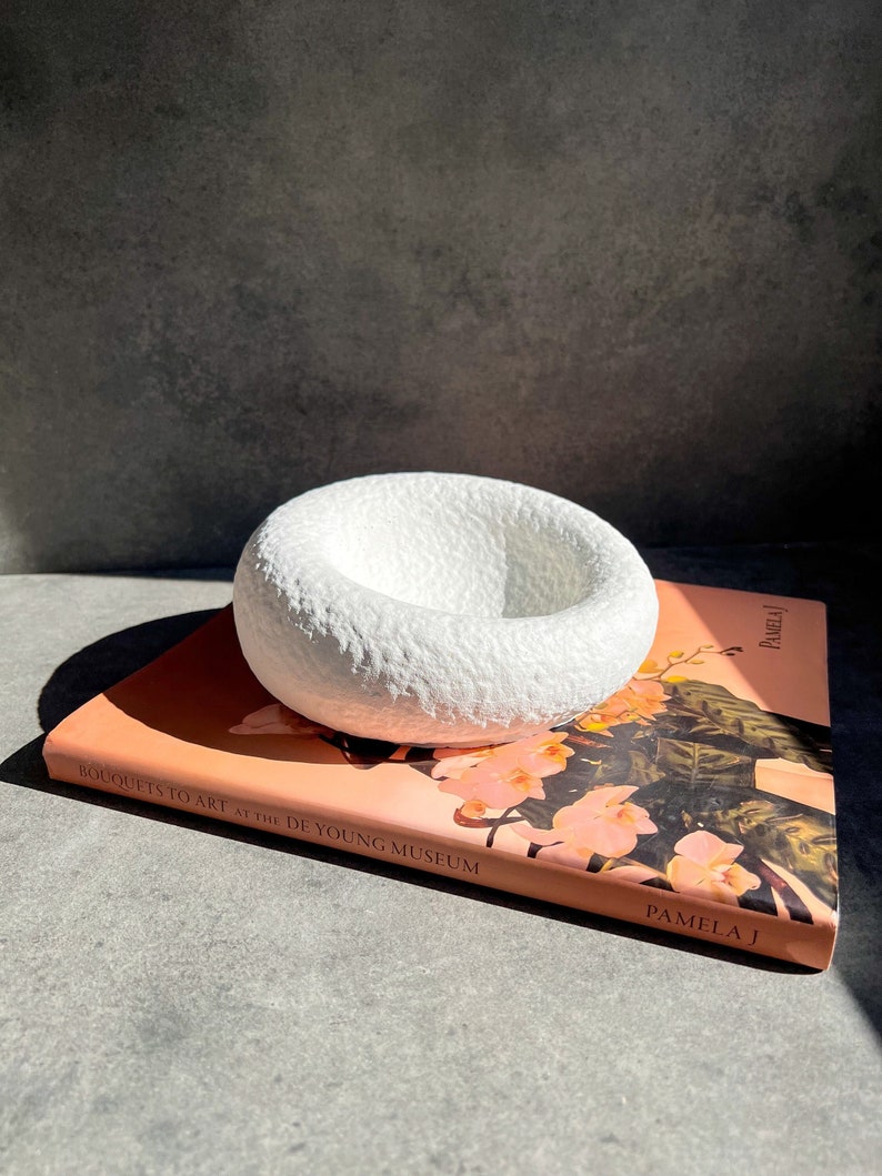 May include: White ceramic bowl with a textured surface, sitting on top of a pink book titled "Bouquets to Art at the de Young Museum" by Pamela J.