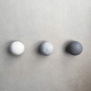 May include: Three round concrete knobs in different shades of gray. The knobs are arranged in a row from left to right, with the lightest shade on the left and the darkest shade on the right.