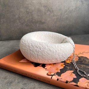 May include: White ceramic bowl with a textured surface, sitting on a book titled "Bouquets to Art at the de Young Museum" by Pamela. A silver chain is draped over the bowl.