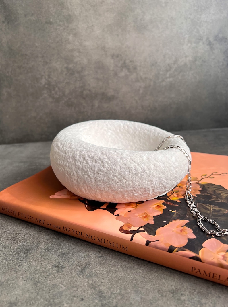 May include: White ceramic bowl with a textured surface, sitting on a book titled "Bouquets to Art at the de Young Museum" by Pamela. A silver chain is draped over the bowl.