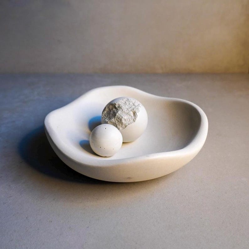 May include: A white, organically shaped bowl contains two spherical objects. One sphere is whole, the other is partially broken, showing a textured interior. The bowl and spheres are a light, neutral colour.