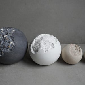 Puede incluir: Cuatro objetos decorativos esf&eacute;ricos de varios tama&ntilde;os y colores. El m&aacute;s grande es gris oscuro, el siguiente es blanco, seguido de una esfera beige, y el m&aacute;s peque&ntilde;o es gris. Cada uno tiene un parche rugoso texturizado.