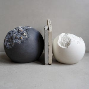 Puede incluir: Naturaleza muerta de tres esculturas abstractas. Una esfera gris oscuro con detalles texturizados se encuentra junto a una esfera blanca con una secci&oacute;n tallada. Una pieza rectangular se interpone entre ellas.