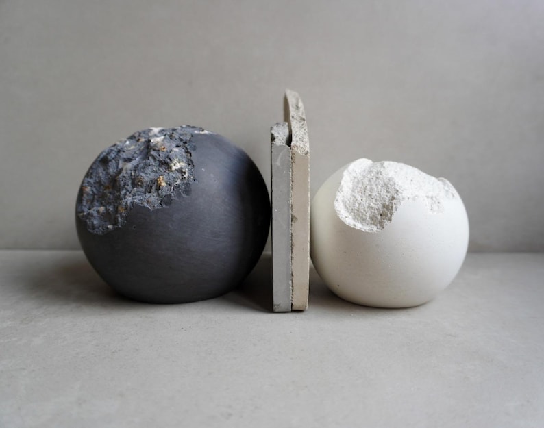 Puede incluir: Naturaleza muerta de tres esculturas abstractas. Una esfera gris oscuro con detalles texturizados se encuentra junto a una esfera blanca con una secci&oacute;n tallada. Una pieza rectangular se interpone entre ellas.