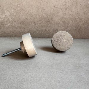 May include: Two concrete cabinet knobs, one with a screw, the other without. The knobs are a light gray color and have a rough texture.