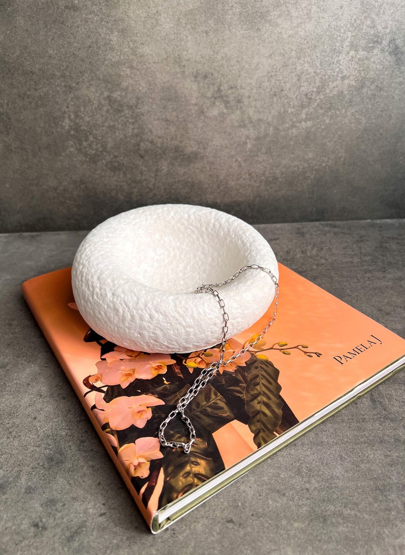 May include: White ceramic hanging planter with a silver chain, resting on a book with a floral design and the text "Pamela J".