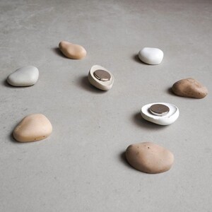 K&ouml;nnte beinhalten: Eine Sammlung von kieselsteinf&ouml;rmigen Magneten in verschiedenen neutralen Farbt&ouml;nen, darunter Beige, Wei&szlig; und Braun. Einige Magnete haben eine flache, vertiefte Fl&auml;che mit einer silbernen Scheibe. Die Magnete liegen auf einer grauen Oberfl&auml;che.