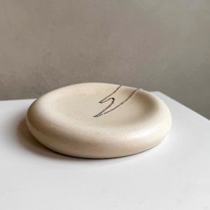May include: A white, round, concrete dish with a silver chain necklace resting inside.