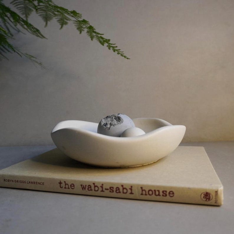 May include: A white, organically shaped ceramic bowl holding two grey decorative spheres and a white sphere, resting on a book titled "the wabi-sabi house". A fern branch is in the background.