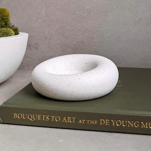 May include: A white, donut-shaped decorative object with a speckled texture. The object sits on a green book with gold lettering that reads "BOUQUETS TO ART AT THE DE YOUNG MU". A white, bowl-shaped object with green moss is partially visible in the background.