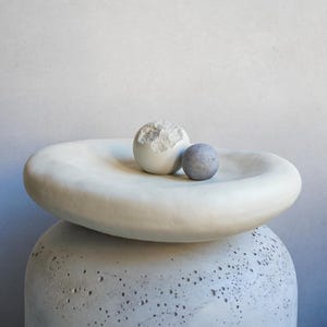 May include: A white, organic-shaped bowl with two spherical objects on top. One sphere is white with a textured surface, and the other is a small, smooth, gray sphere. The bowl rests on a textured, gray, rounded base.
