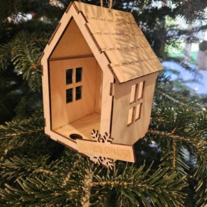 May include: A wooden house-shaped Christmas ornament with a shingled roof and cut-out windows. The ornament has the text "ELVES ONLY!" on a snowflake-shaped sign. It hangs from a natural twine string, displayed on a Christmas tree.