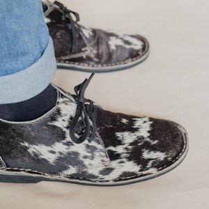 Puede incluir: Un par de zapatos de cordones de cuero de vaca negro y blanco. Los zapatos tienen una suela de goma negra y un cord&oacute;n negro.