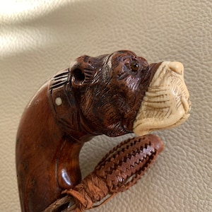 Puede incluir: Un bastón de madera con una cabeza de bulldog tallada. La cabeza del bulldog está hecha de madera oscura con un hocico de marfil blanco. El bastón tiene una correa de cuero.