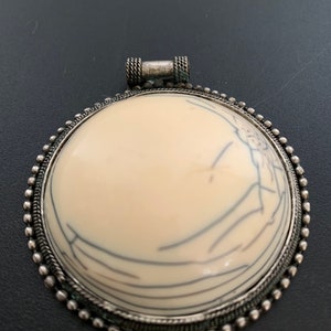 Peut inclure: Un pendentif rond avec une incrustation d'os blanc et un cadre en métal argenté. Le cadre est bordé de perles et comporte une boucle en haut pour l'accrocher.