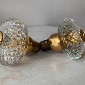 May include: Two vintage glass door knobs with a diamond pattern and gold-colored metal accents. The knobs have a classic design, with a clear, faceted glass body and a decorative gold-toned center.