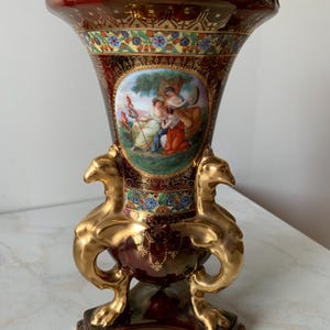 Può includere: Un vaso decorativo in porcellana con una smaltatura bordeaux profondo, decorato con filigrana dorata e motivi floreali. Il centro presenta un medaglione circolare con una scena dipinta di tre figure, fiancheggiato da due manici dorati a forma di grifone.