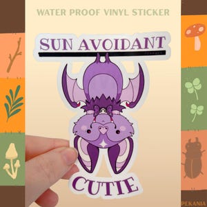 May include: A purple bat sticker with the words "SUN AVOIDANT" and "CUTIE". The bat has large ears and wings, and is hanging upside down. The sticker is water proof vinyl.