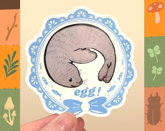 May include: A water-proof vinyl sticker featuring a gray seal in a blue and white decorative frame. The seal is curled up with the word "egg!" below. The sticker has a bow at the bottom and the text "WATER PROOF VINYL STICKER" at the top.