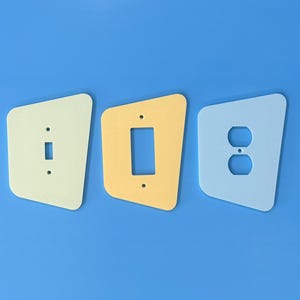 May include: Three uniquely shaped light switch covers in cream, yellow, and light blue. The covers are for a toggle switch, a rectangular outlet, and a double outlet. The background is a solid blue.
