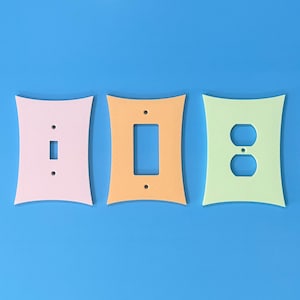 May include: Three decorative light switch covers in pastel colors: pink, orange, and green. Each cover has a unique cutout for a switch or outlet. The covers are arranged against a solid blue background.