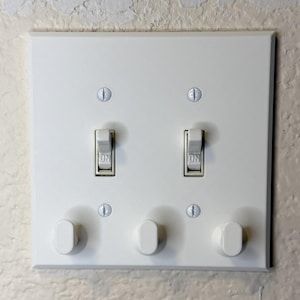 May include: A white electrical switch plate with two toggle switches and three oval-shaped light switch dimmers. The switches are in the "ON" position. The plate is mounted on a textured wall.