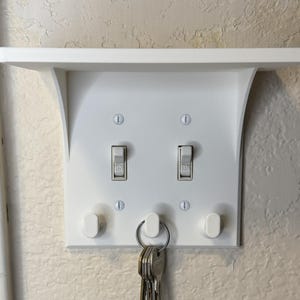 May include: White wall-mounted key holder with a shelf. Features two light switch-style toggles and two key hooks. A set of keys hangs from the bottom. The holder is mounted on a textured beige wall.
