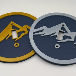 May include: Two round wall decor pieces with a mountain silhouette design. One is gold and the other is gray, both set against a dark blue background. Each has a cutout for a light switch and a small hole.