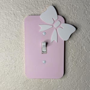 May include: A light pink light switch cover with a white bow accent. The bow is attached to the top of the cover. The switch is in the center of the cover, with two screws at the top and bottom.