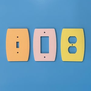 May include: Three colorful light switch covers in shades of orange, pink, and yellow. The covers are oval-shaped with cutouts for a switch, a rocker switch, and a standard outlet. The background is a solid blue.